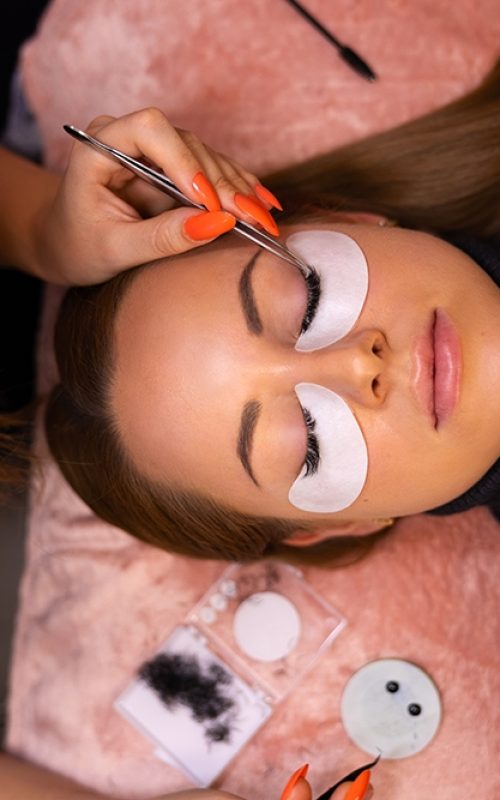 Overhead view of beautiful female client going through eyelash extension procedure while cosmetologist using tweezers